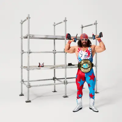 A muscular man dressed as wrestler Macho Man with a championship belt, holding an 18V 2-in-1 impact driver in each hand, standing in front of a metal scaffolding structure.