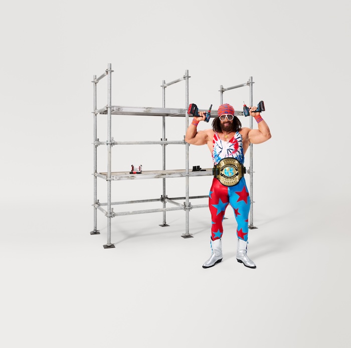 A muscular man dressed as wrestler Macho Man with a championship belt, holding an 18V 2-in-1 impact driver in each hand, standing in front of a metal scaffolding structure
